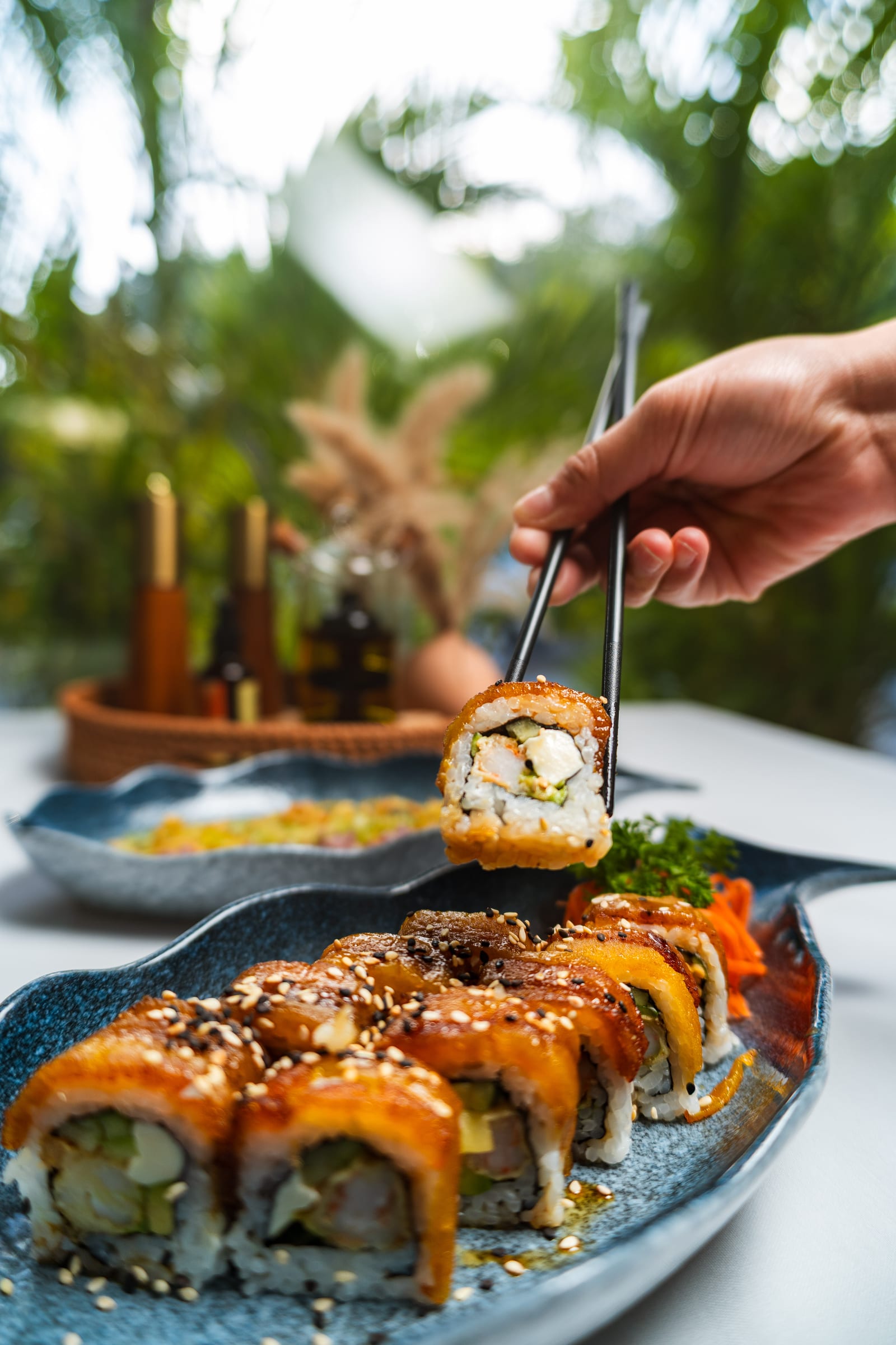 Chopsticks lifting a piece of MERO 18's plantain-topped Plátano con Queso sushi roll