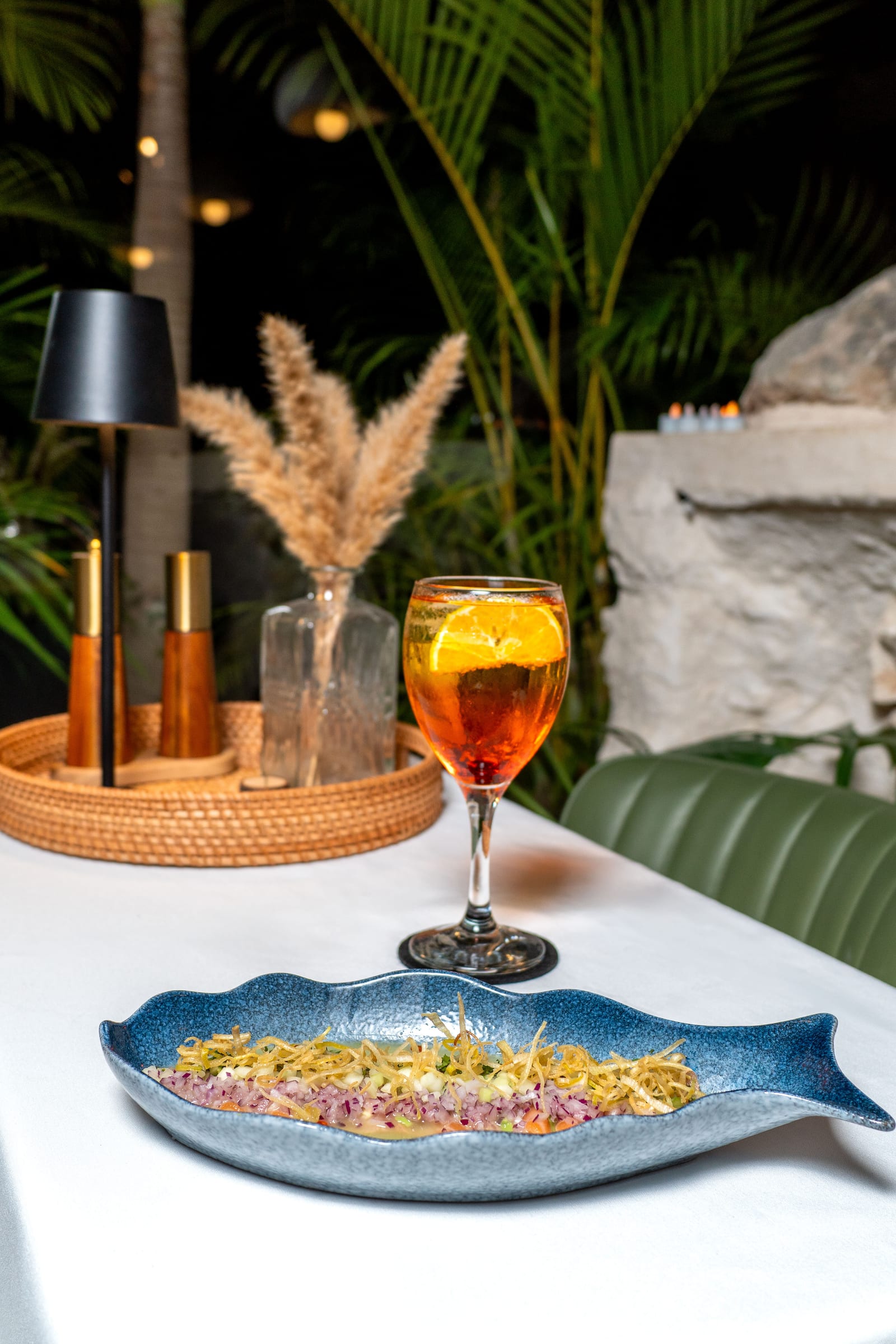 Ceviche on a fish-shaped blue plate with a spritz cocktail on a night table among palms at MERO 18