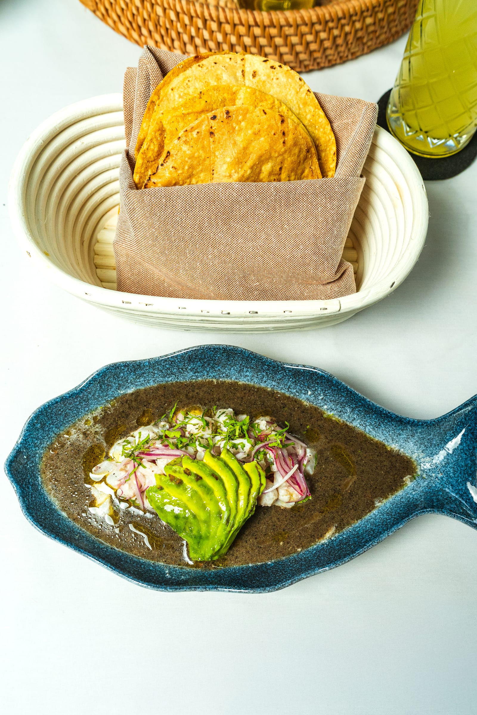 Aguachile negro con abanico de aguacate y cebolla morada en plato con forma de pez, con canasta de tostadas en MERO 18 Cancún