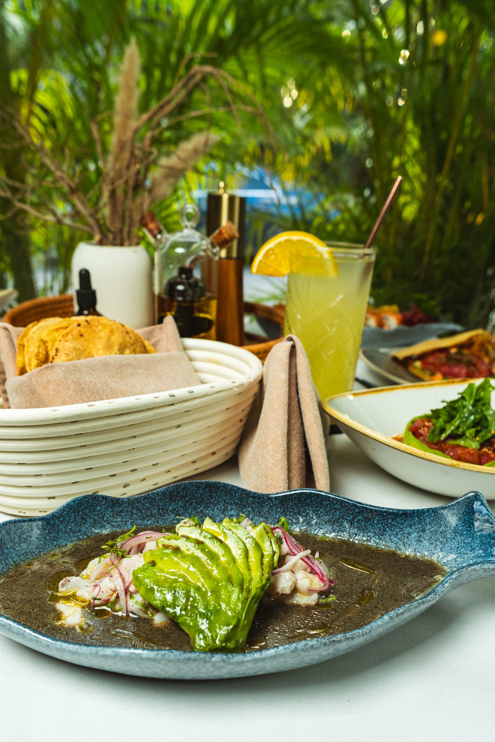 Mesa de MERO 18 con aguachile negro en plato azul con forma de pez, tostadas, limonada y tostada de atún en el patio selvático