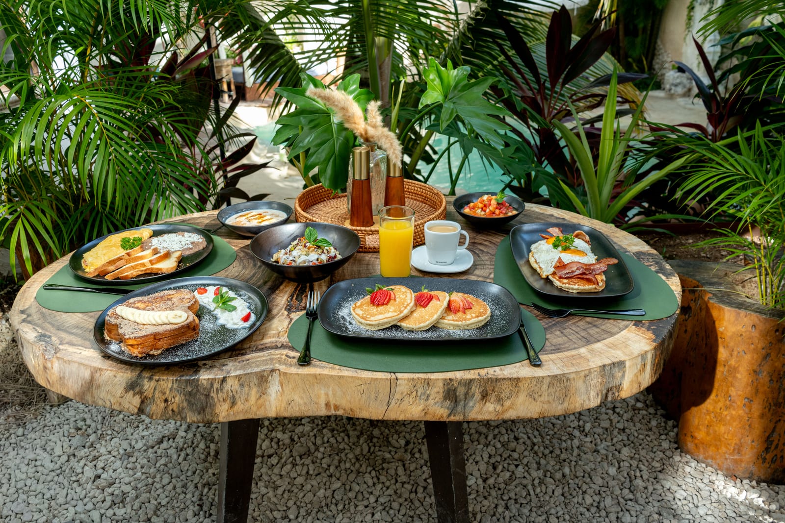 Mesa de desayuno de MERO 18 junto a la alberca cenote: hotcakes, pan francés, omelette y jugos en el jardín selvático