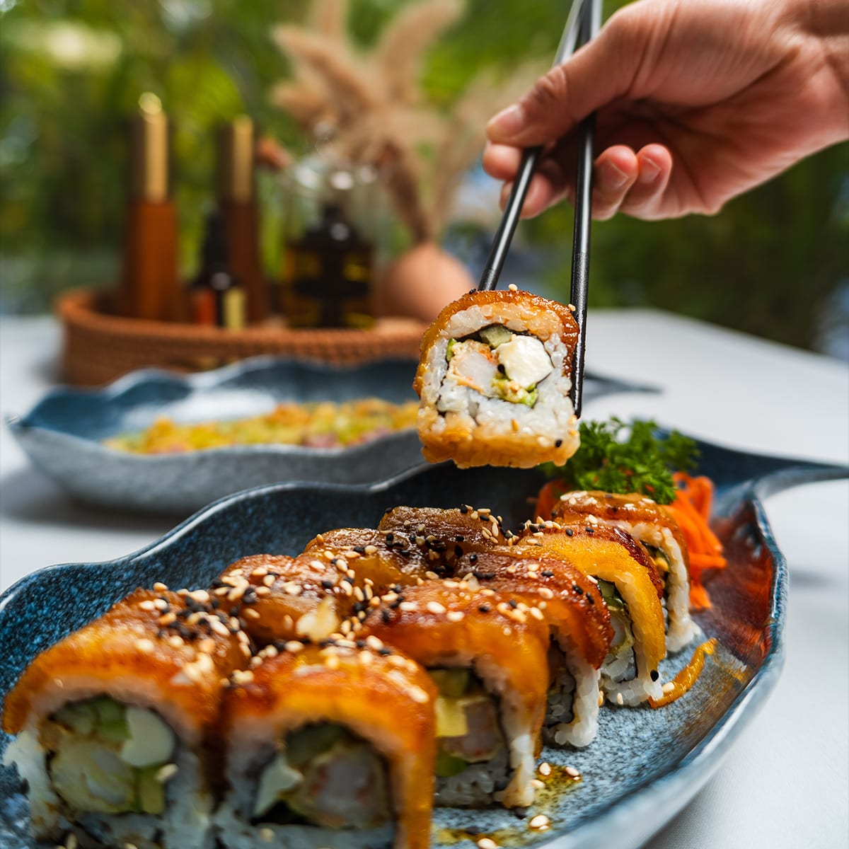 Palillos levantando un rollo de sushi glaseado sobre plato azul en MERO 18, la foto de sushi más fácil de lograr en un restaurante del Centro de Cancún